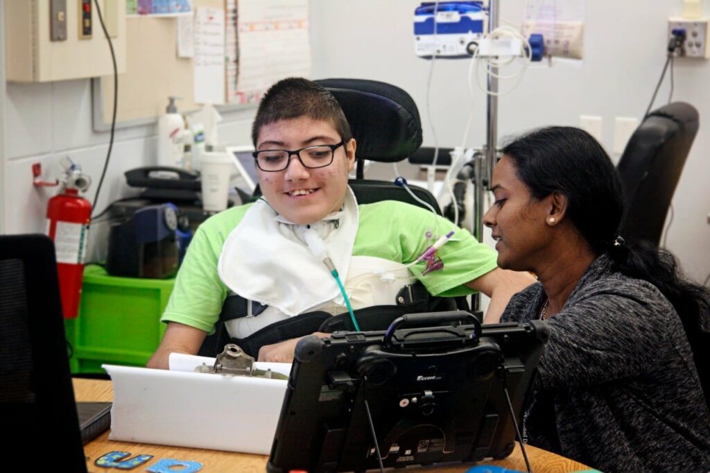 Student-with-AAC-Device - Building Wings An educator works with a student in a wheelchair with complex needs.