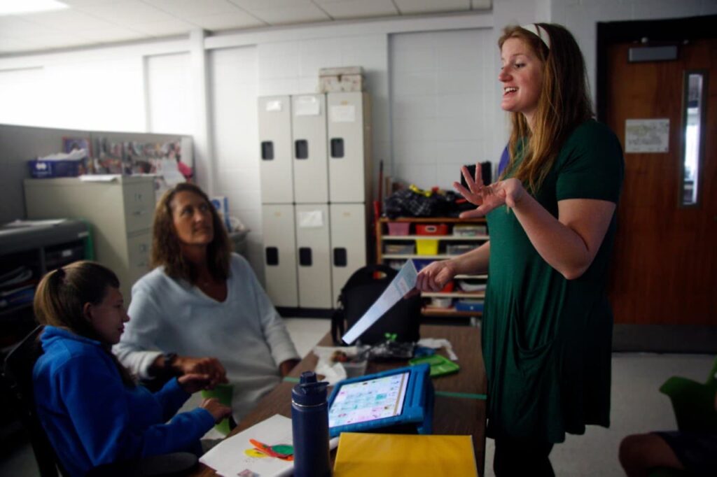 Abbey-Lynch-Header - Building Wings Abbey Lynch speaks to two people sitting at desks in a classroom in MacArthur Middle School.