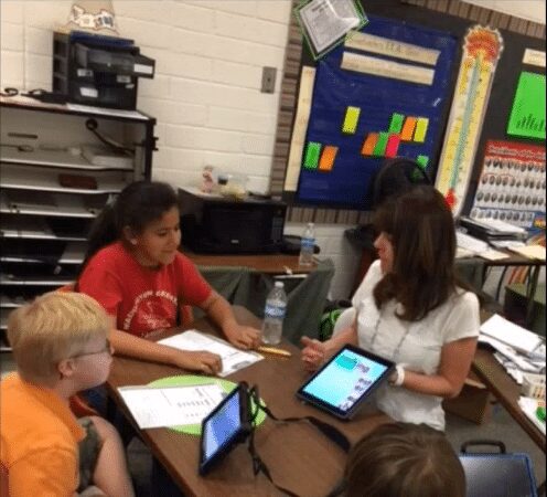 Word-Study-JMJ - Building Wings Teacher engages in a word study with three students at a desk, in a classroom.
