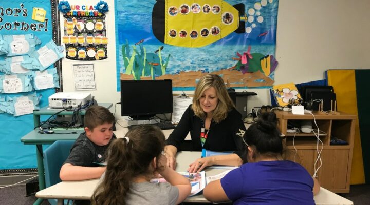 A teacher leads a reading lesson with three students at a desk in a classroom, with marine life creations on the walls.