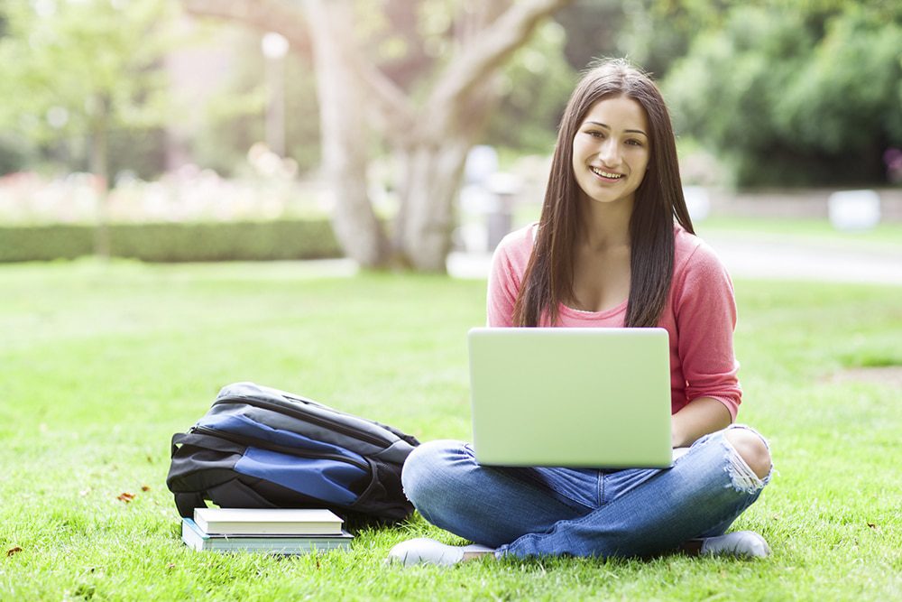 Article-2b-1000w - Building Wings A girl sitting crosslegged outdoors smiles at the camera with a laptop in her lap and a backpack next to her on the ground.