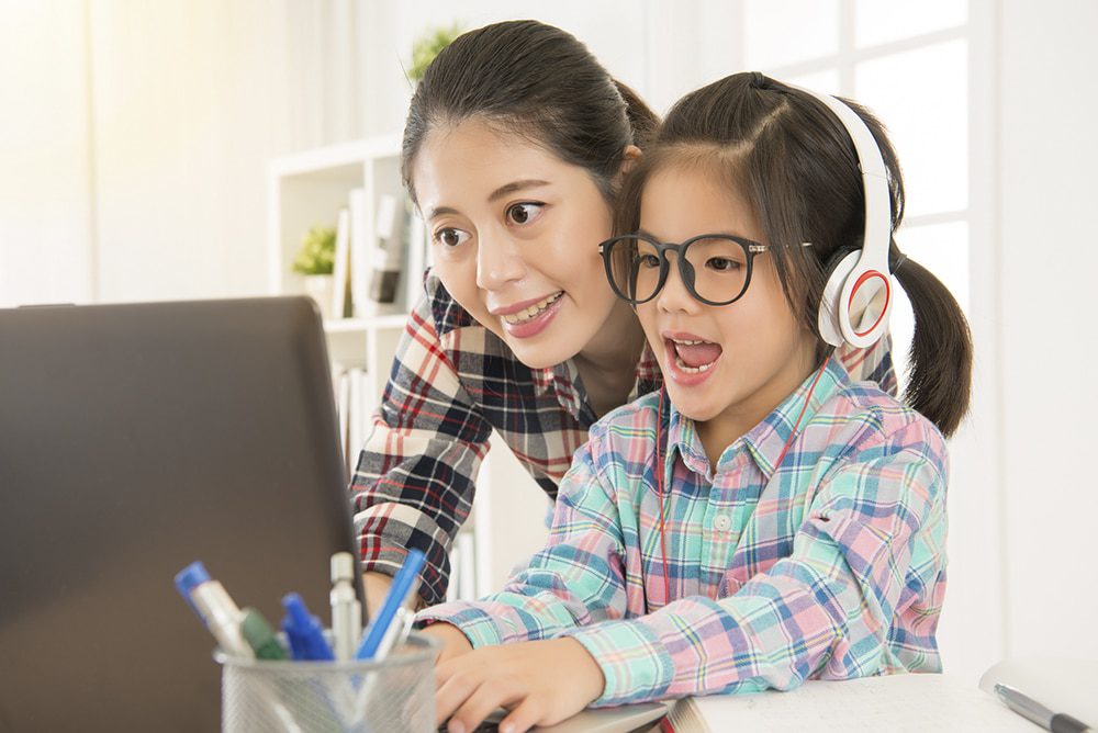 Article-2d-1000w - Building Wings A mother smiling with her daughter who is wearing headphones and glasses while working at a computer in a home