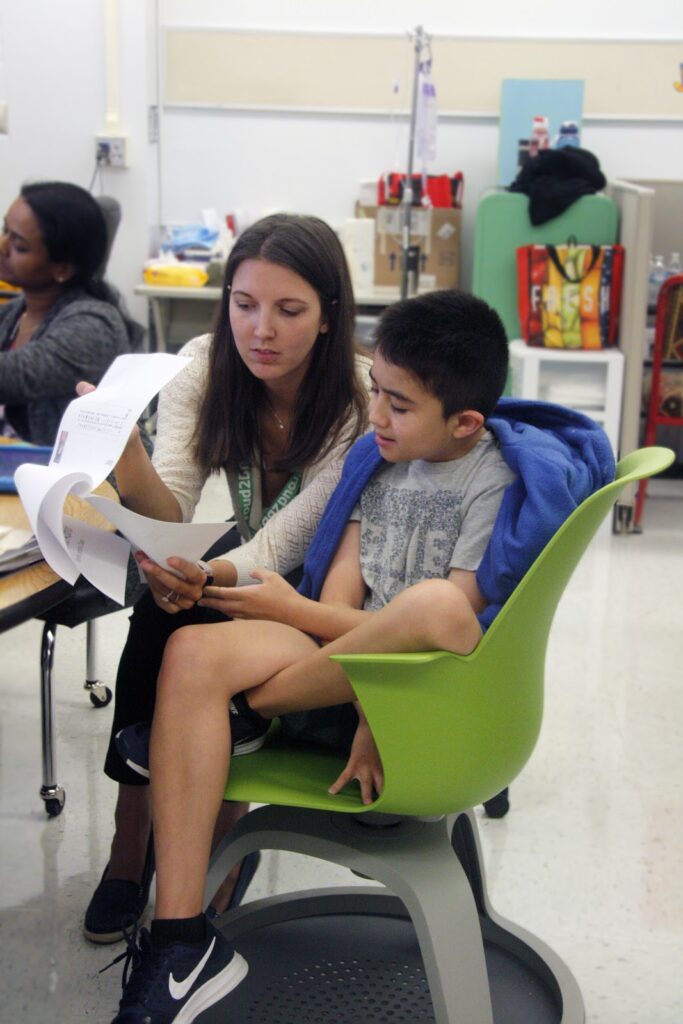 student-with-teacher - Building Wings A teacher reads to a student sitting in a green chair in a classroom.