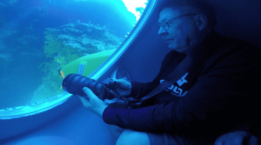 Don Johnston holds a camera up to the window of submarine to take a photograph of the underwater sea terrain that can be seen through the window.