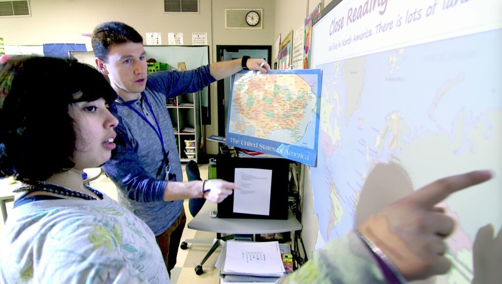 Erlind Lacy holds up a physical map of the U.S. while a student points to a digital map of the world on a screen, in a classroom.