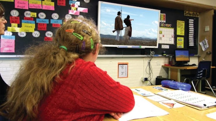Student wearing a red shirt in a classroom watching a Readtopia video lesson on a smart board.