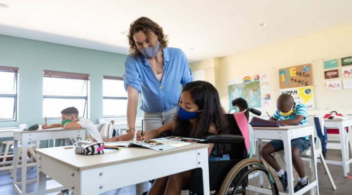 A teacher wearing a mask work helps a student who is reading a Readtopia lesson in a classroom, with other students working in the background.