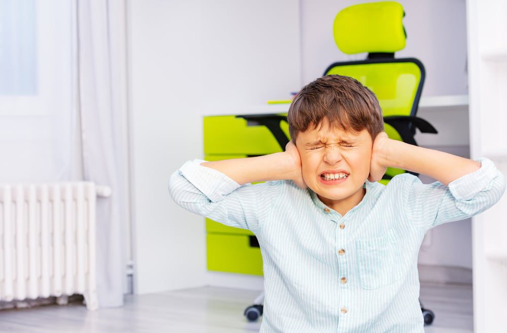 Depositphotos_410470288_S - Building Wings A young child holds his hands to his ears with an uncomfortable look on his face and his eyes closed.