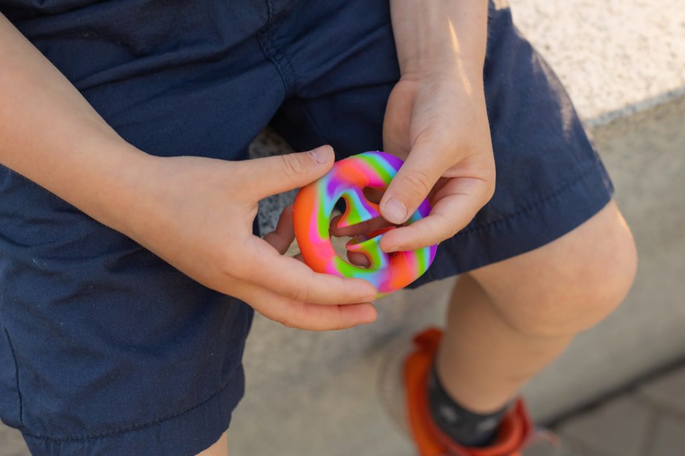 Depositphotos_478524984_S - Building Wings A young child holds a colorful stimming toy, while sitting a cement stoop outdoors.