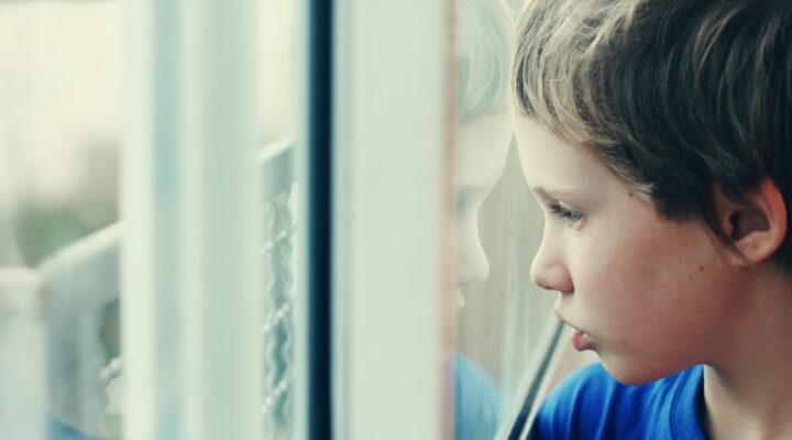 A young child in a blue shirt stares longingly out a window.