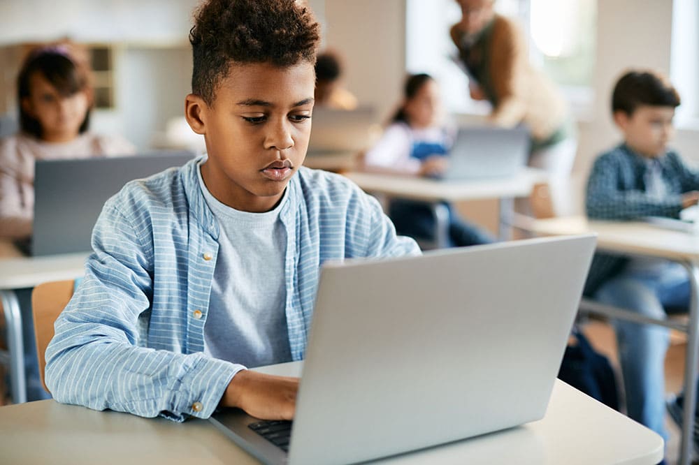 student-using-laptop-classroom - Building Wings Student uses a laptop at a desk, in a classroom, with other students and a teacher blurred in the background.
