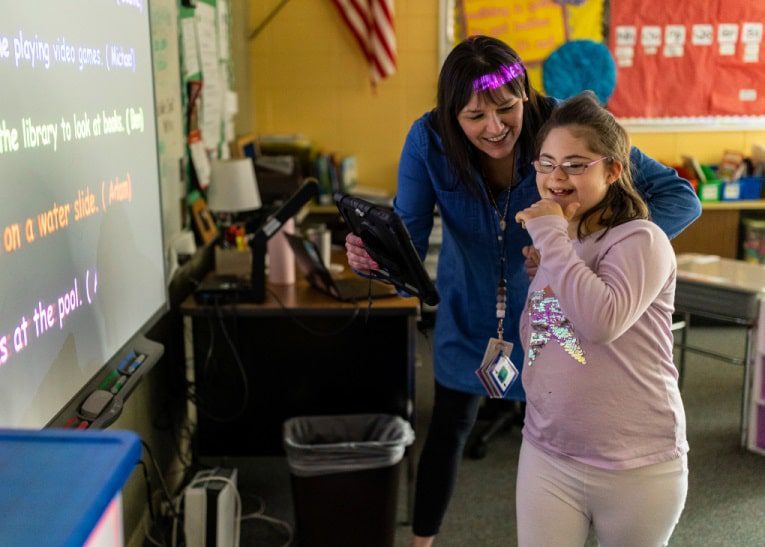 Naperville2022_Prairie Elementary_IMN_273-min - Building Wings A teacher smiles at a laughing student, while the student presents in front of a smart board in a classroom.