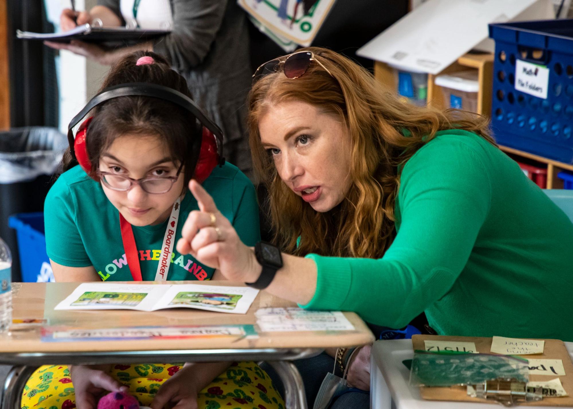 Classroom image 3 - Building Wings In a classroom, a teacher wearing a green shirt, points something while working with a student sitting at a desk.
