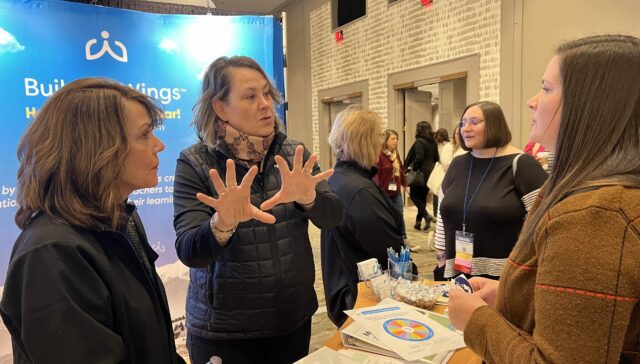 CTG 2022 Building Wings and ReadtopiaGO - Building Wings Maureen Donnelly uses her hands to describe something to two women in front of a large Building Wings poster at a conference, with other people standing, and having conversations in the backgrounds.