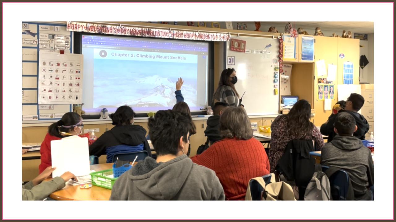 BW Evidence of Impact v2.pptx (3) - Building Wings Classroom setting taken from the rear of the room showing the backs of learners who are looking at a screen showing a chapter from a Readtopia Unit next to an educator at the front of the room.