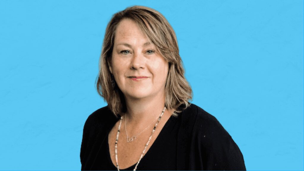Maureen Donnelly headshot on Blue Background - Building Wings A headshot of a woman, Maureen Donnelly, with shoulder-length brown hair, wearing a black shirt and necklace, against a blue background.
