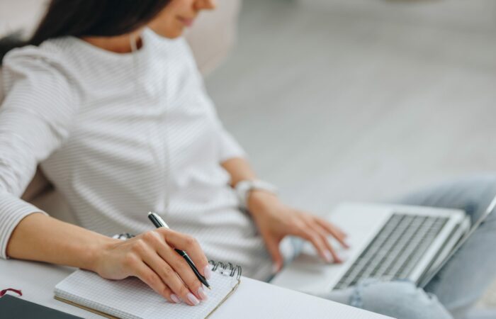 Virtual coaching for special educators - Building Wings A woman learning remotely with one hand on the keyboard of a laptop computer and another hand holding a pen near a pad of paper.