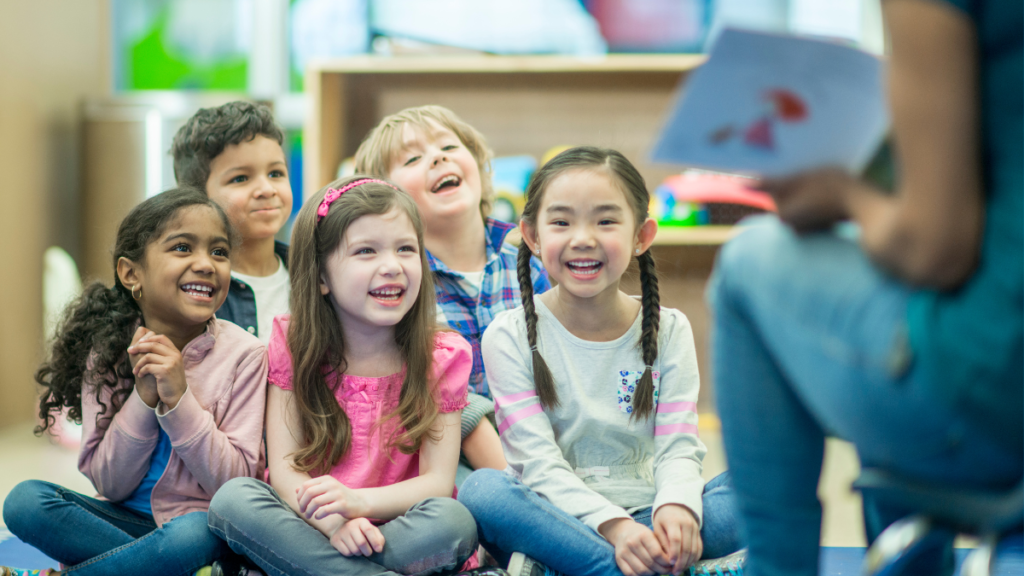 Shared Reading in a Classroom - Building Wings A group of young students sitting on a classroom floor facing a teacher who is reading out loud from a book.