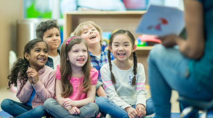 A group of young students sitting on a classroom floor facing a teacher who is reading out loud from a book.