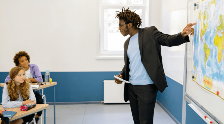 A male teacher in front of a classroom pointing to a map while students are looking towards the map - using options to connect one of the strategies for teaching current events