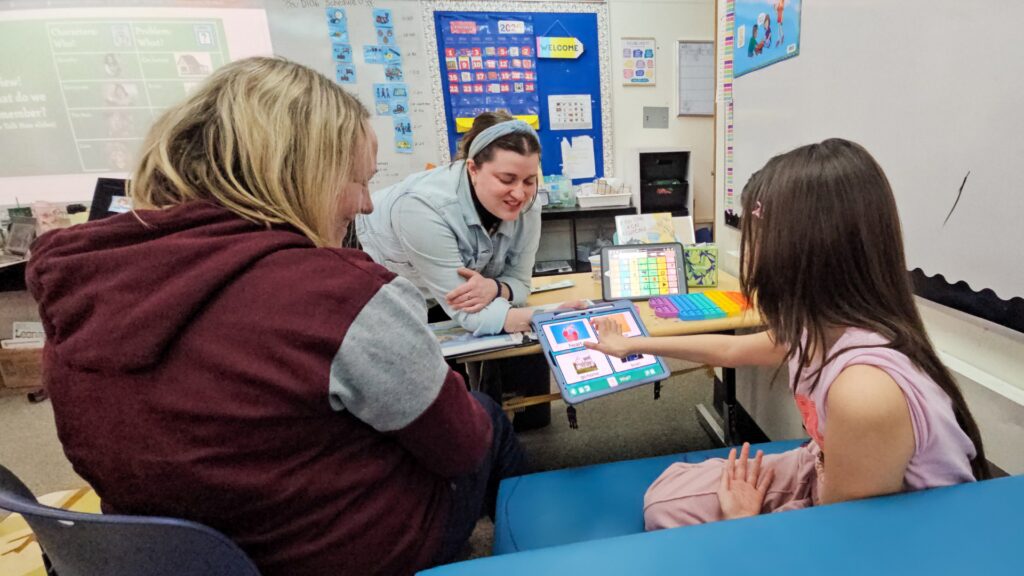 Teacher guides two students in a classroom as they use tablets for learning or communication.