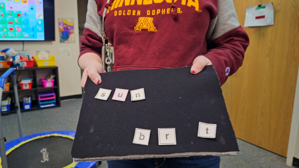Person wearing a maroon Minnesota hoodie holds a black foam board with letter tiles spelling 'sun' on top row and 'brt' on bottom row in a classroom.