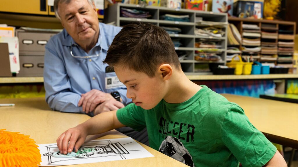 Young boy in a green shirt colors a page at a table while an adult supervisor watches in a classroom setting.