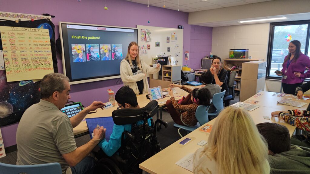 Teacher guides a pattern-learning activity for a diverse group of children and adults at a long table; a screen shows 'Finish the pattern!'