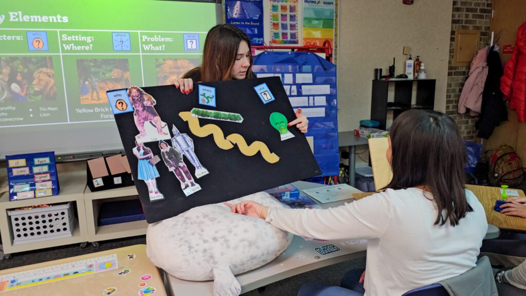 Student presents a collage board with Wizard of Oz characters (Dorothy, Scarecrow, Tin Man, Lion) in a classroom setting. Behind, a green project screen shows learning prompts.