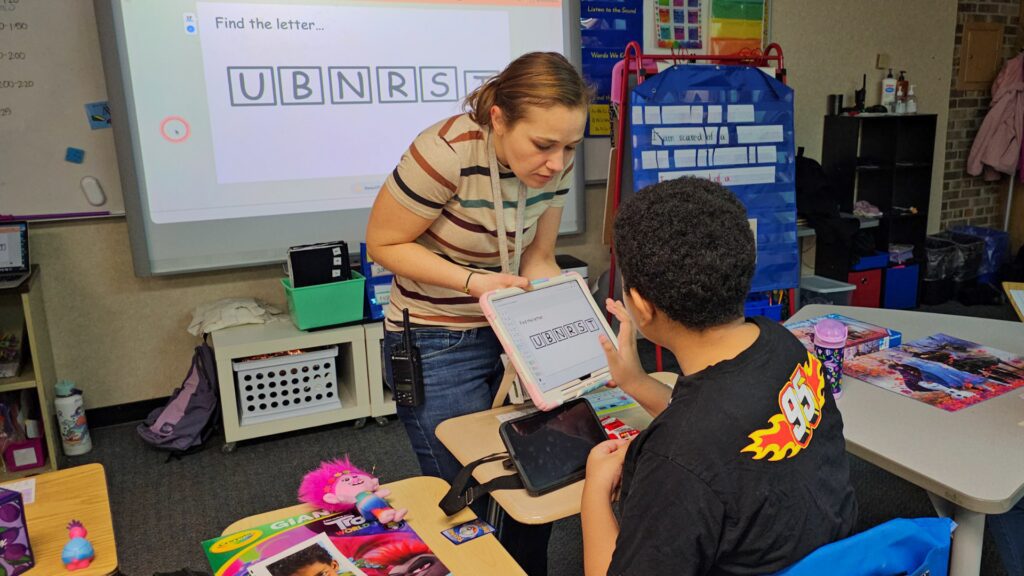 Teacher assists a student with a tablet during a classroom activity; screen shows letters U B N R S T in tile blocks on the tablet.