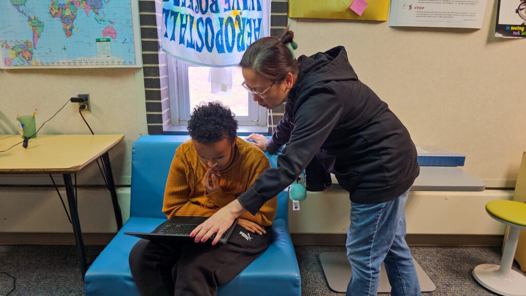 Teacher leans over a student on a blue chair, helping with a laptop in a classroom. World map and posters on the wall behind them.