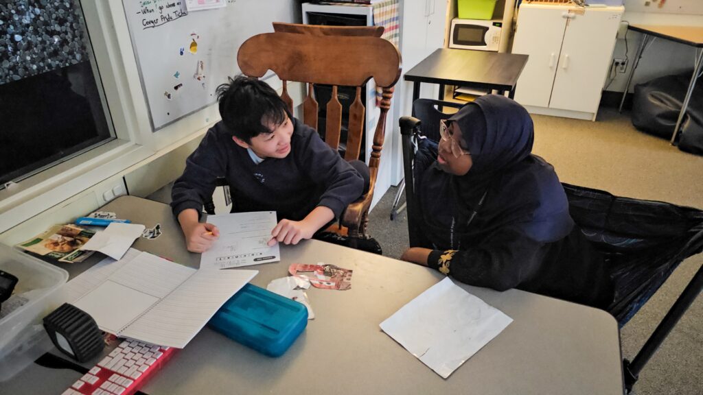 Student sits at a table doing worksheets while a teacher or aide in a hijab looks on and smiles in a classroom setting.