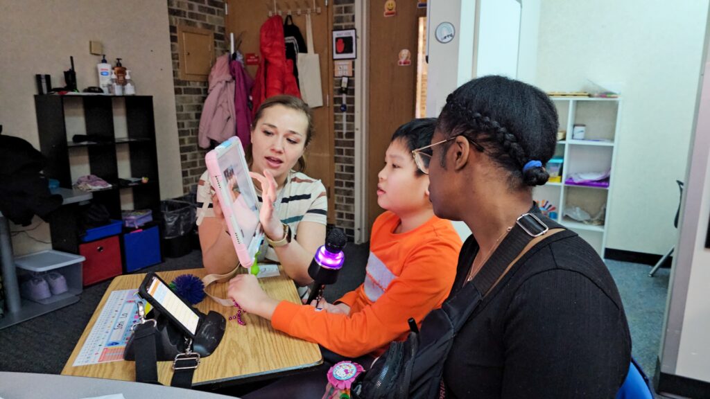 Woman demonstrates a tablet to a young boy as another woman watches in a classroom setting.