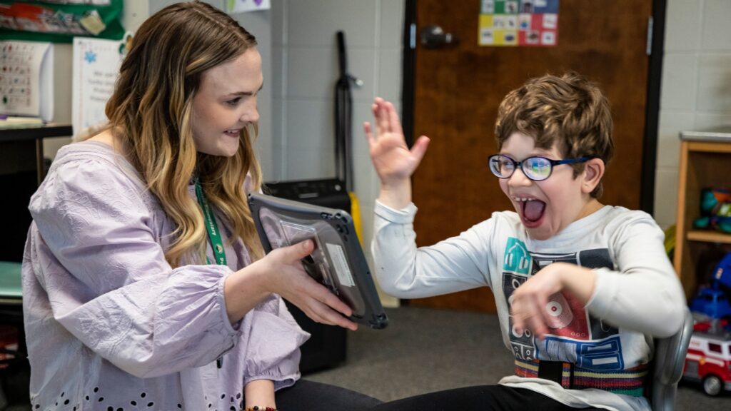 Young boy with glasses high-fives a smiling female teacher who holds a tablet in a classroom setting.