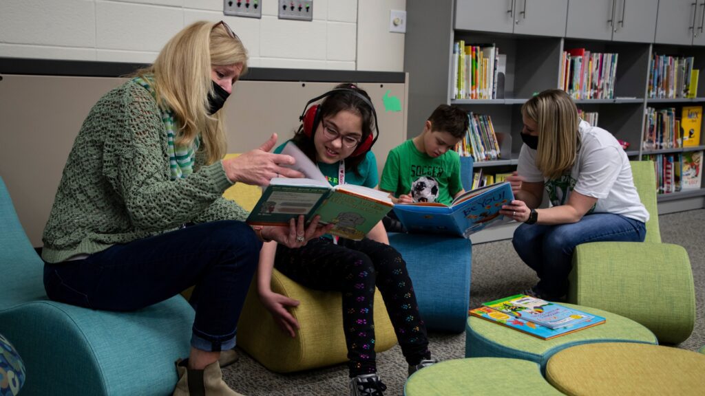 Adults and children sit on colorful lounge chairs in a library, reading books together and smiling at the pages.