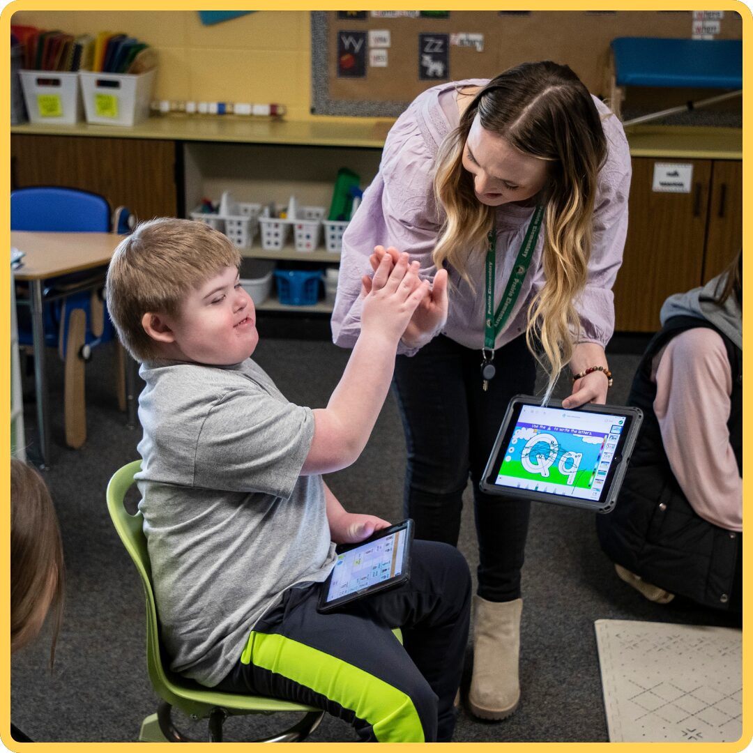 Teacher high-fiving a seated student in a classroom while both use tablets for learning.