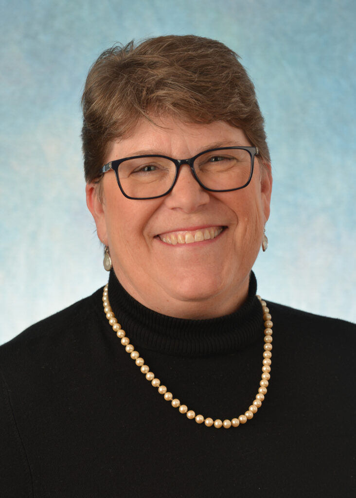 Woman smiling with a black top, short brown hair, and gold necklace on a blue background