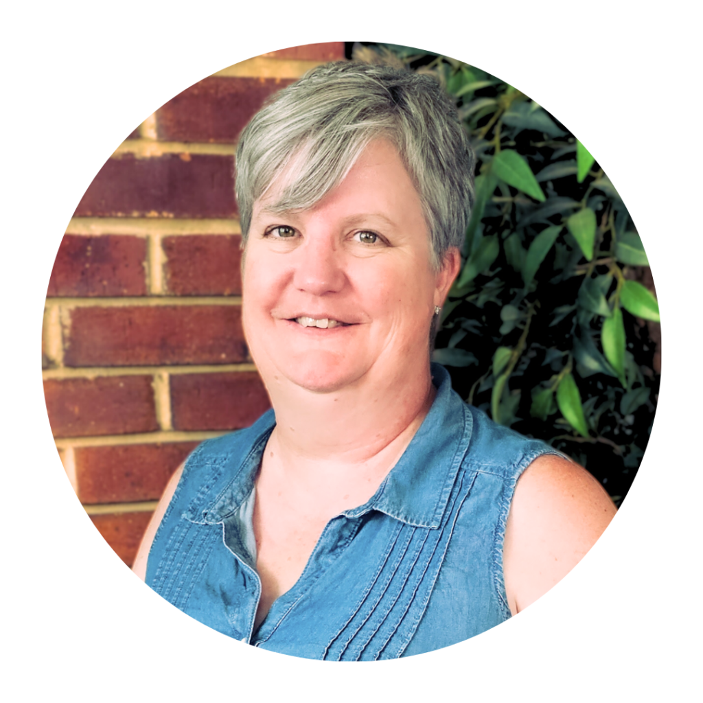 Portrait of a smiling woman with short gray hair wearing a denim sleeveless shirt, with brick wall and green foliage in the background.
