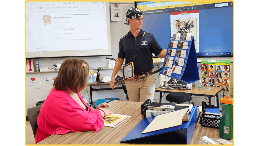 Teacher leading a Readtopia Treasure Island literacy lesson using a classroom display and character communication supports while a student follows along at a table.